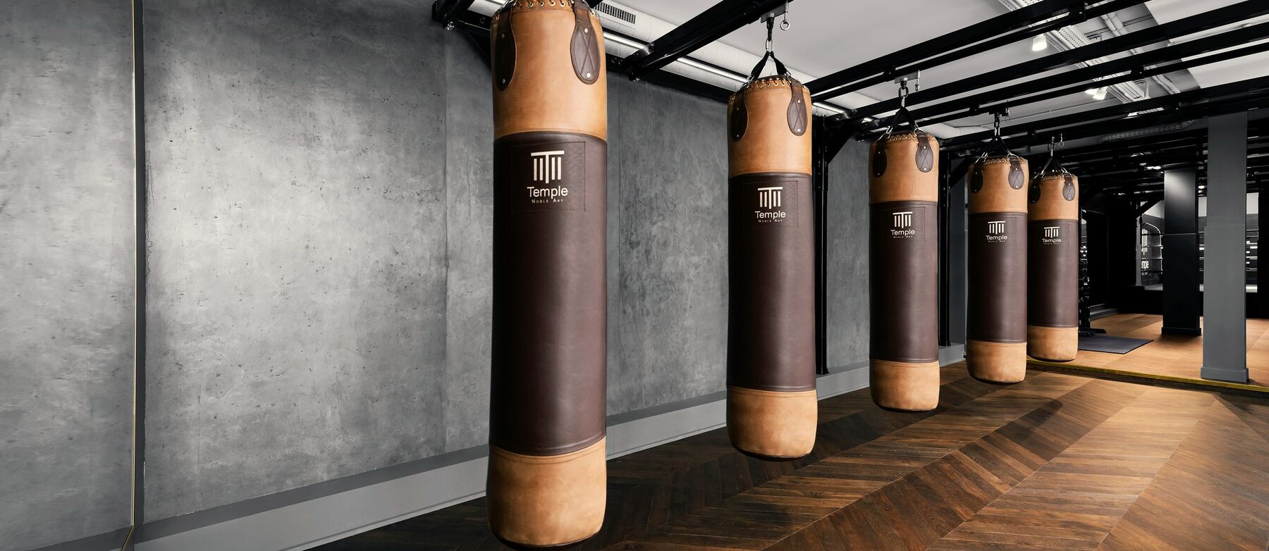 A row of leather punching bags in a gym with concrete walls and a parquet floor